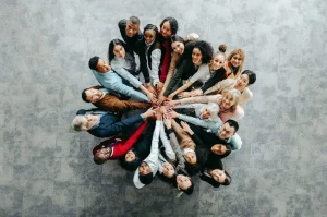 A diverse group of people standing in a circle, looking up at the camera, with their hands joined together in the center.