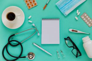 Medical supplies including a stethoscope, pills, a blister pack of capsules, syringe, face mask, notepad, digital thermometer, a cup of coffee, and eyeglasses arranged on a turquoise background.