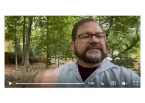 A video thumbnail featuring a man with glasses and a beard speaking outdoors in front of a background of green trees. A video playback bar and controls are visible at the bottom of the frame.